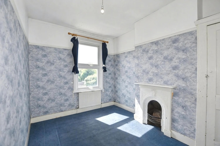 A bedroom with its original cast iron fireplace intact, blue patterned wall paper above the dado and blue carpet throughout