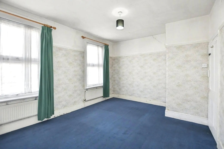 A second bedroom with two windows, green curtains and pale floral wall paper, cracked plaster above the picture rail