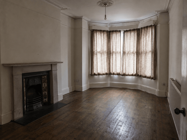 The empty front room of a period house, curtains drawn across the bay window, bare floorboards and a cast iron fireplace